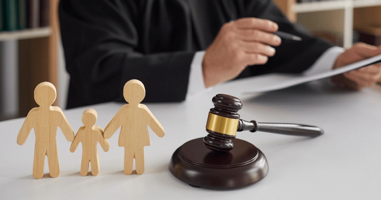 family-law-solicitors-7275099_1280 (1) Ein Richter in schwarzer Robe sitzt an einem Tisch. Im Vordergrund steht ein Holzhammer (Richterhammer), daneben drei Holzfiguren – zwei größere Figuren (Eltern) und eine kleinere Figur (Kind) – die eine Familie symbolisieren.